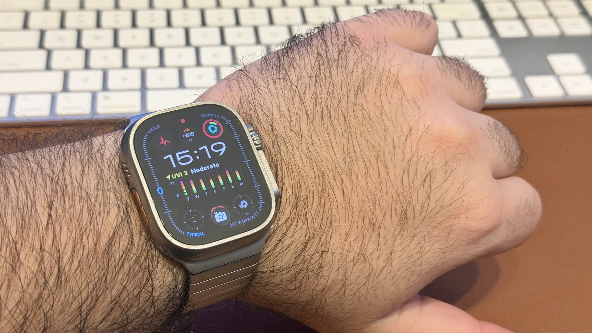 Apple Watch Ultra (1st generation) with a titanium link bracelet on a wrist, in front of a white keyboard and a brown desk mat